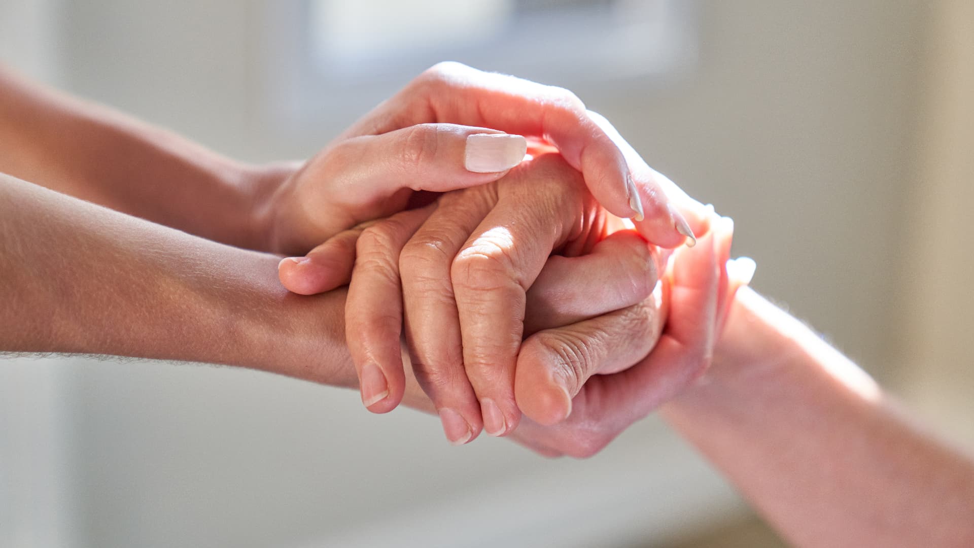A younger person gently holding an older person's hands in a bright, calm room
